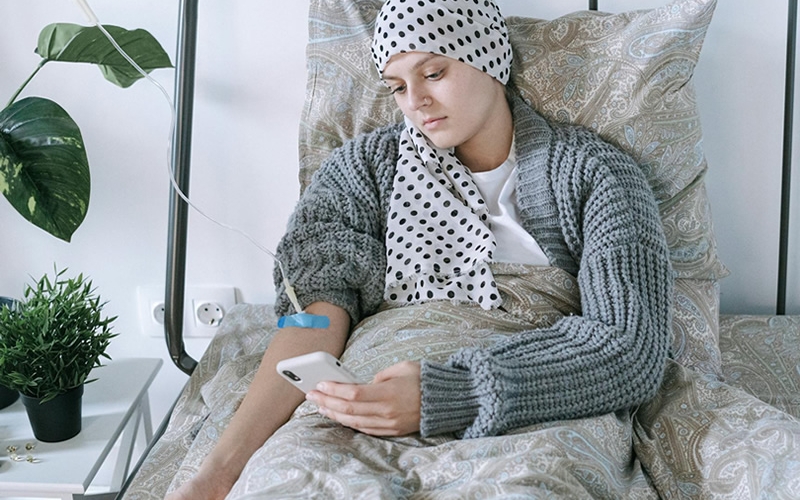 Woman in gray sweater and head scarf looking at phone while receiving an IV treatment sitting in bed with green plants on bedside table.