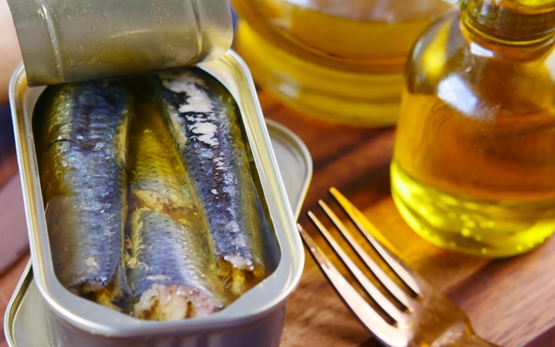 Open tin of sardines on a wood table with a fork and bottle of oil next to it.