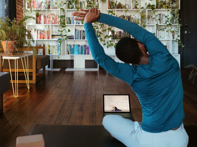 Person seated on mat on floor doing stretching exercises. Wall of books and plants in background.