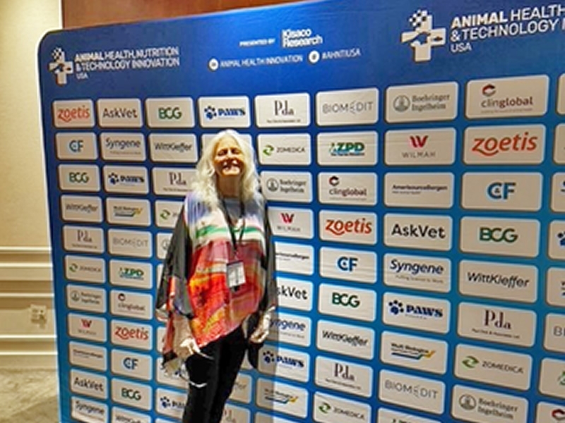 Margo Roman standing in front of a large sign with logos of companies that supported the 2024 Animal Health, Nutrition, and Technology Innovation Conference.