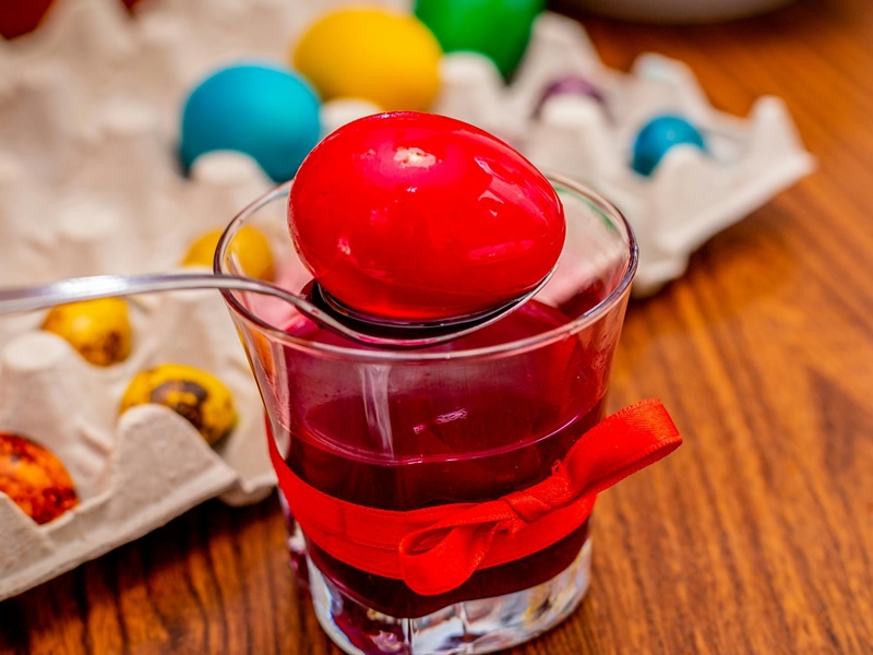 Egg being dyed bright red in foreground; egg carton of different color eggs in background.