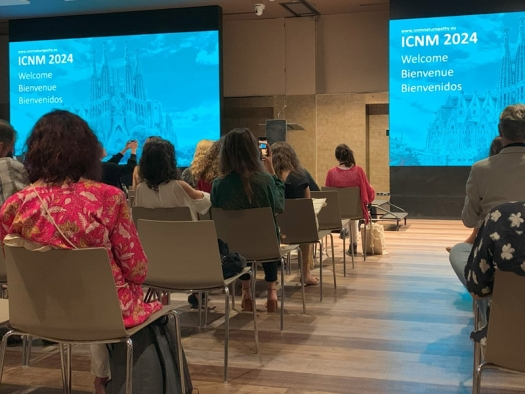 Large conference room with two large screens at the front which say "ICNM 2024" and people seated in chairs waiting for the speaker.