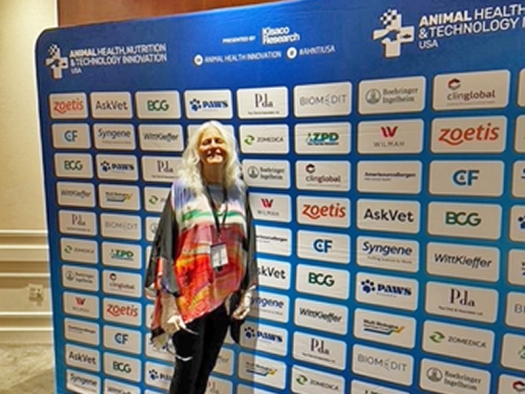 Margo Roman standing in front of a large sign with logos of companies that supported the 2024 Animal Health, Nutrition, and Technology Innovation Conference.