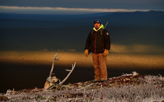 Man in heavy jacket and gloves standing on ground with frost-covered grasses